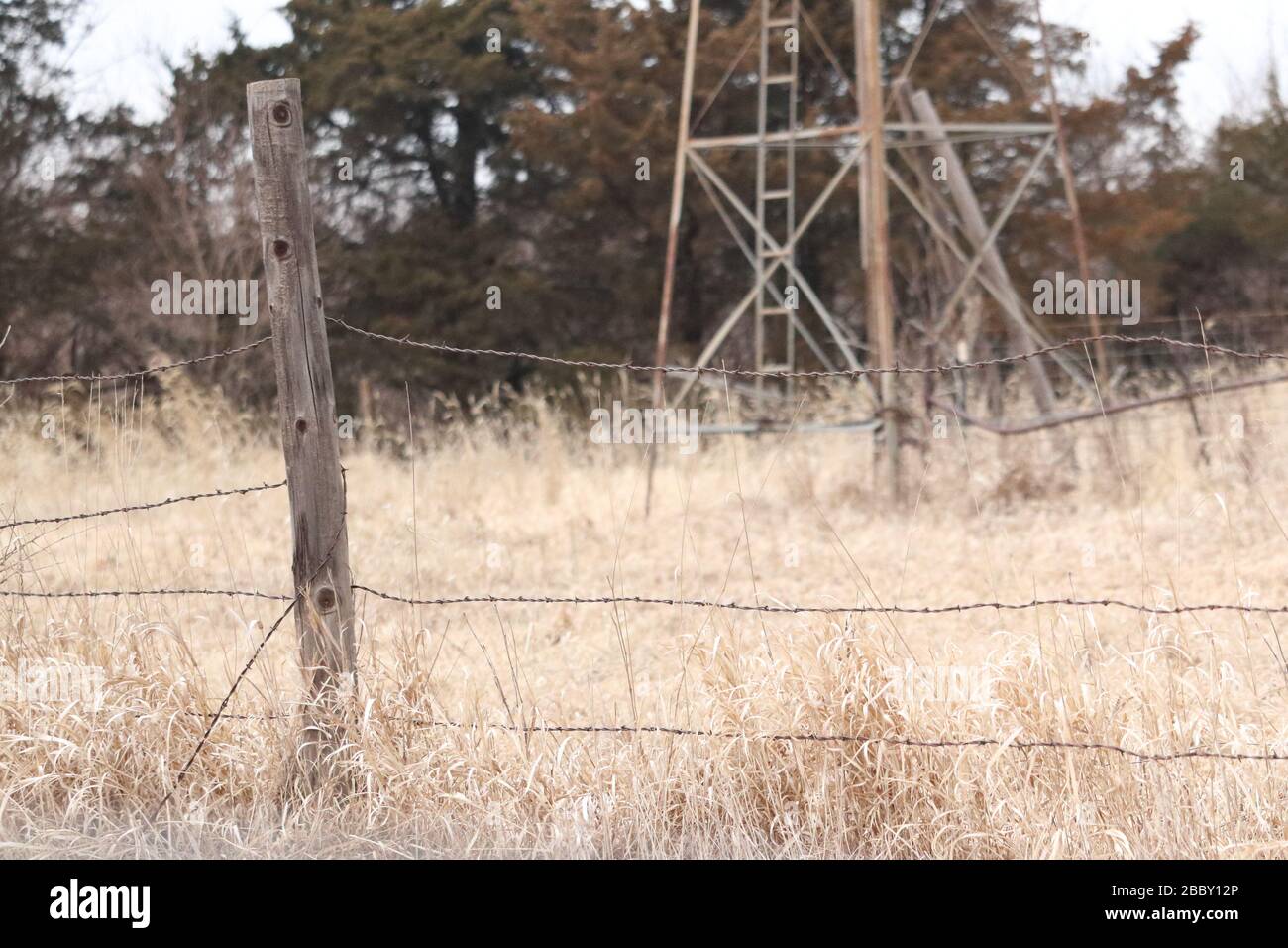 Una chiusura di un vecchio palo di recinzione in legno e filo spinato in un campo di erba asciutta con un vecchio mulino a vento Foto Stock