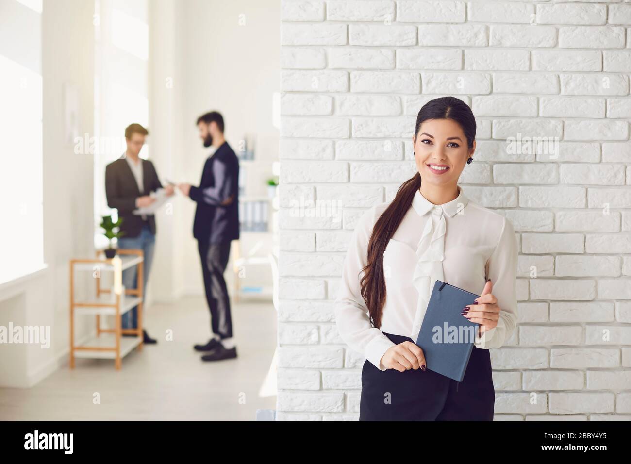 Donna d'affari latina ispanica sorridente mentre si trova in un ufficio bianco contro un muro bianco. Foto Stock