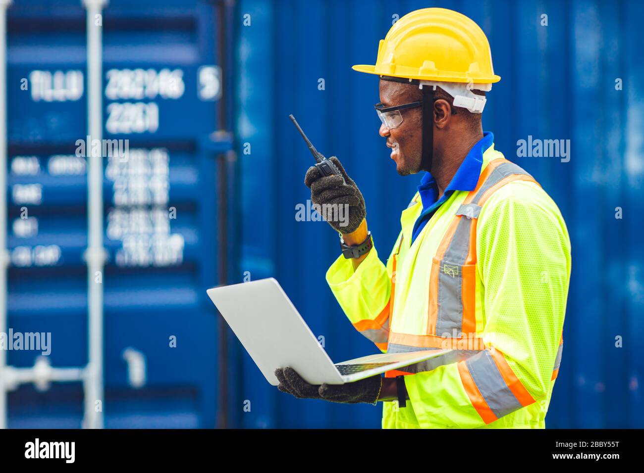 Black African Happy Worker che lavora nella comunicazione logistica utilizzando radio e laptop per controllare i container di carico al porto di carico per i camion per l'esportazione A. Foto Stock