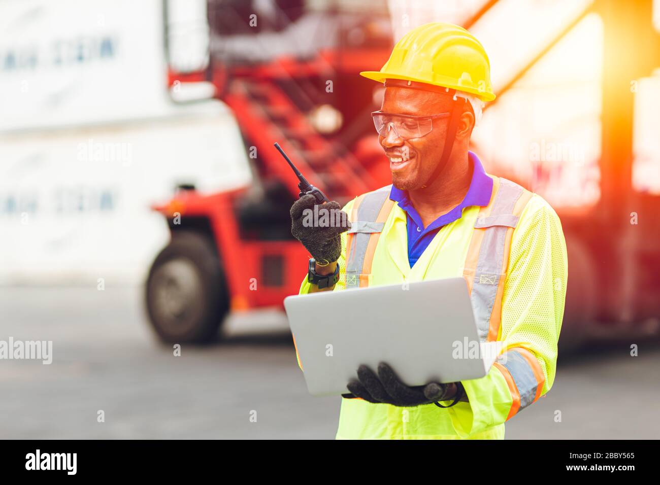 Black African Happy Worker che lavora nella comunicazione logistica utilizzando radio e laptop per controllare i container di carico al porto di carico per i camion per l'esportazione A. Foto Stock