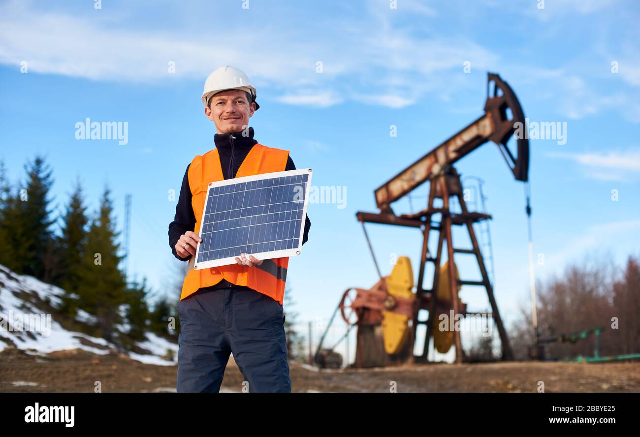 Uomo d'affari sorridente in gilet arancione e casco bianco che tiene mini pannello solare in calda giornata di sole, sullo sfondo il jack pompa e cielo blu, concetto di fonte alternativa di energia Foto Stock