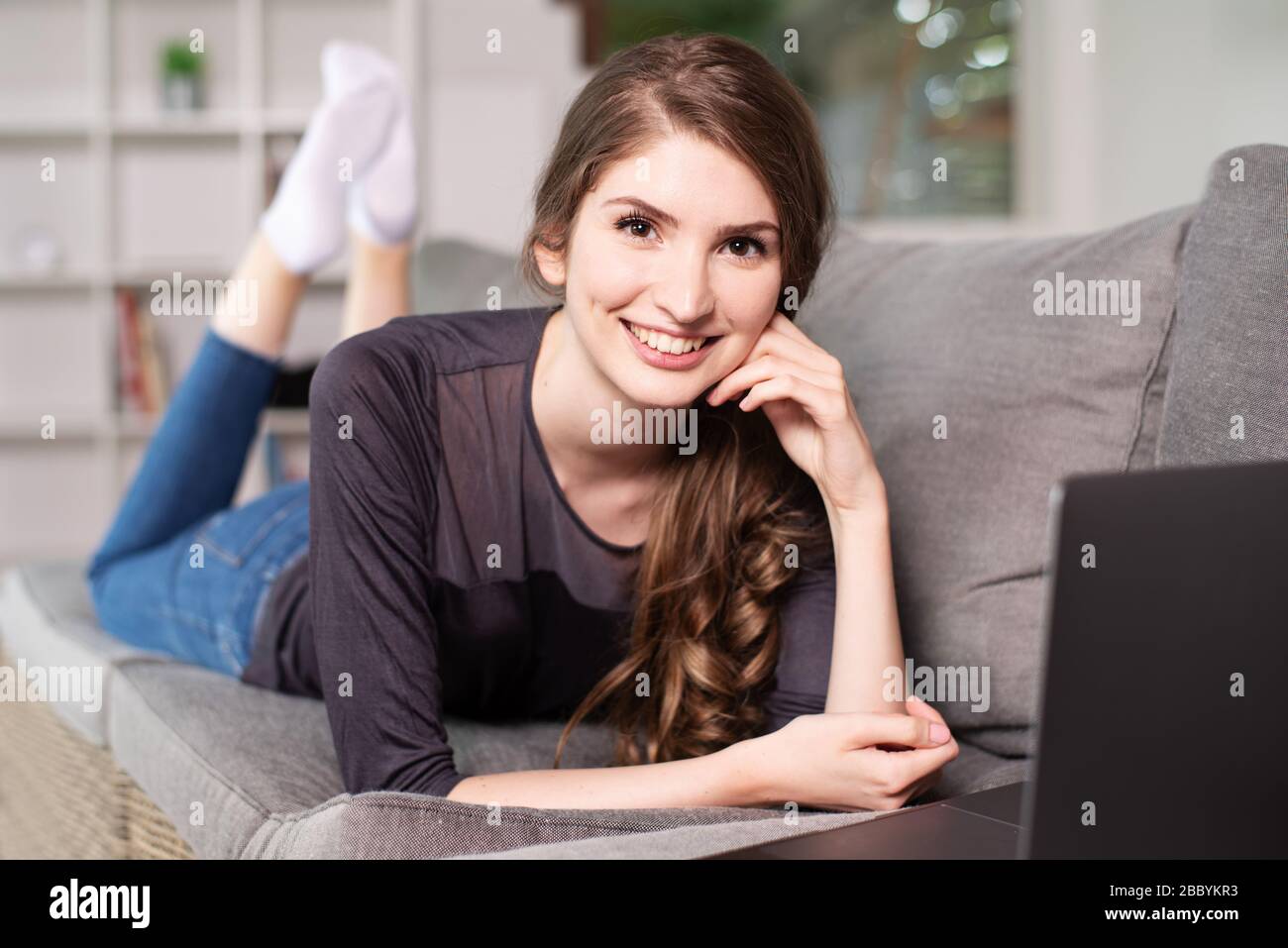 Bella giovane donna sdraiata sul divano con un laptop e sorridente simpateticamente alla macchina fotografica Foto Stock