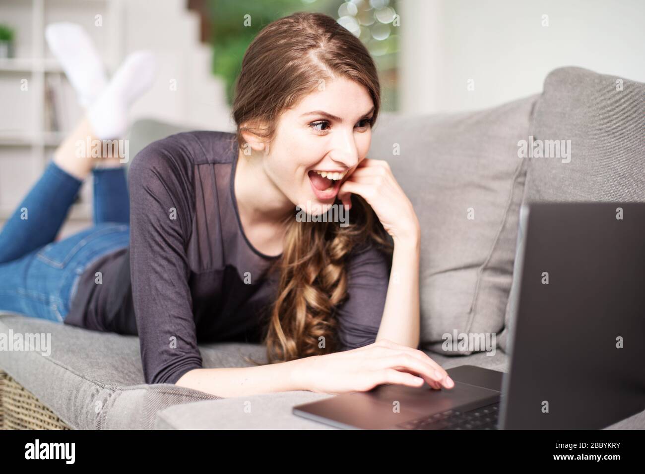 Bella giovane donna sdraiata sul divano con un laptop e sorridente simpateticamente alla macchina fotografica Foto Stock