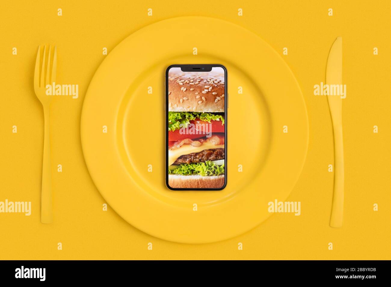 Ordinare cibo online, consegna cibo. Chiama e porta il cibo a casa. Smartphone con hamburger sullo schermo, sdraiato su un piatto, un coltello e una forcella. Giallo. La Foto Stock
