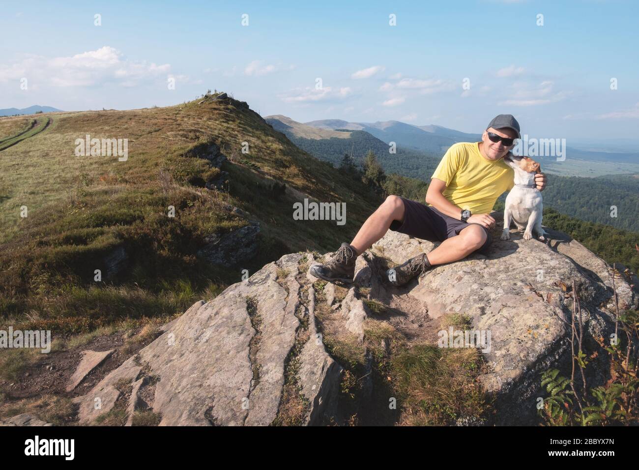 Solo turista seduto sul bordo della scogliera con cane bianco sullo sfondo di un incredibile paesaggio di montagna. Giornata di sole e cielo blu Foto Stock