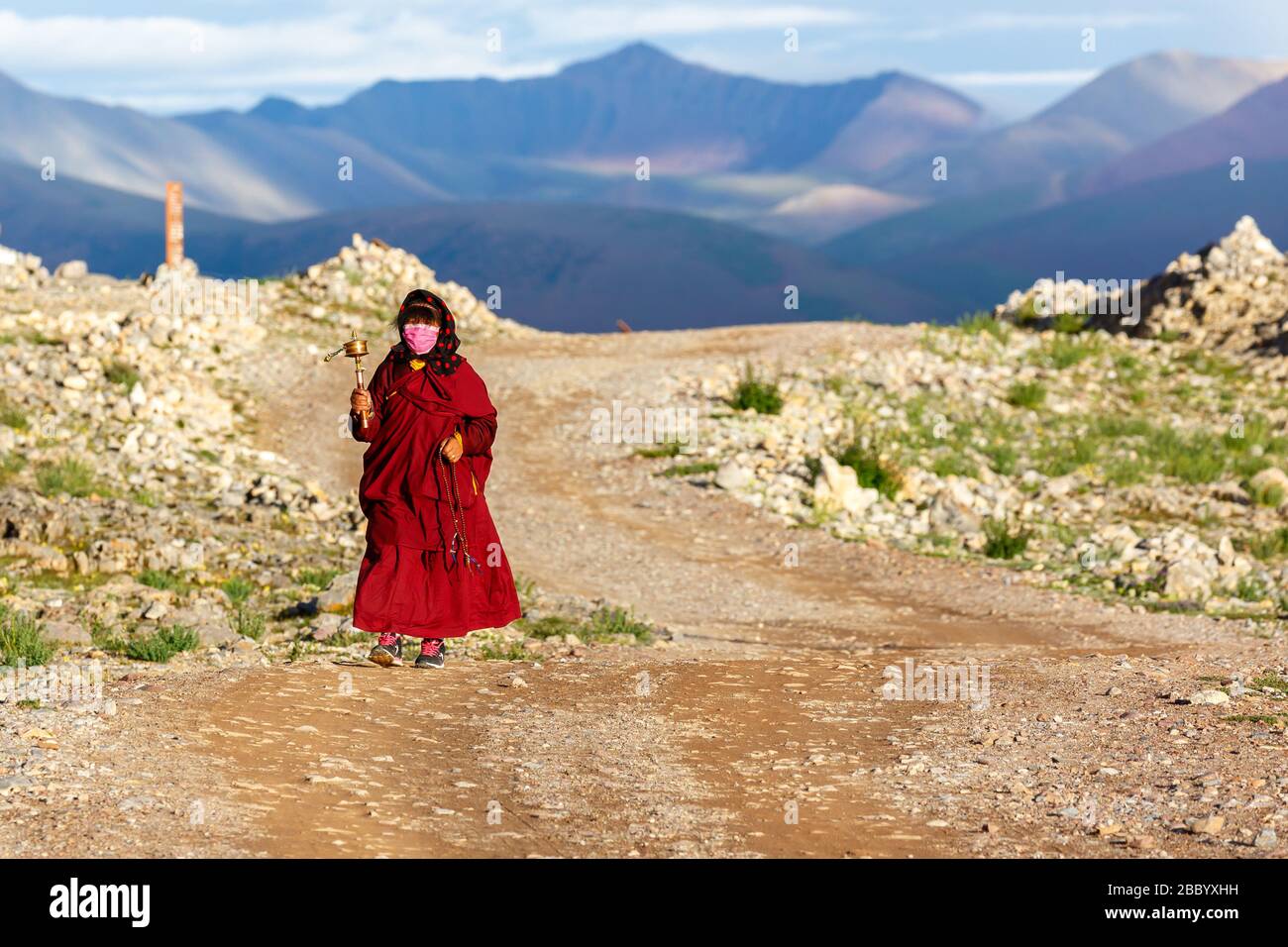 Pellegrino femminile in una tovagola rossa che cammina il Kora. Sta oscillando una ruota di preghiera della mano nella sua mano destra. Nelle montagne posteriori dell'altopiano tibetano. Foto Stock