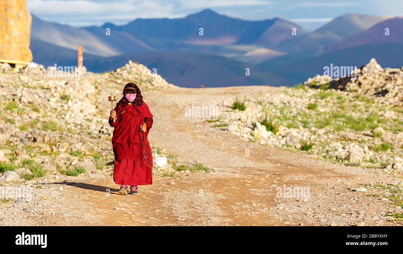 Pellegrino (donna) con accappatoio rosso e ruota di preghiera a mano che cammina il tradizionale Kora al Lago Nam Tso. Montagne dell'altopiano tibetano sullo sfondo. Foto Stock