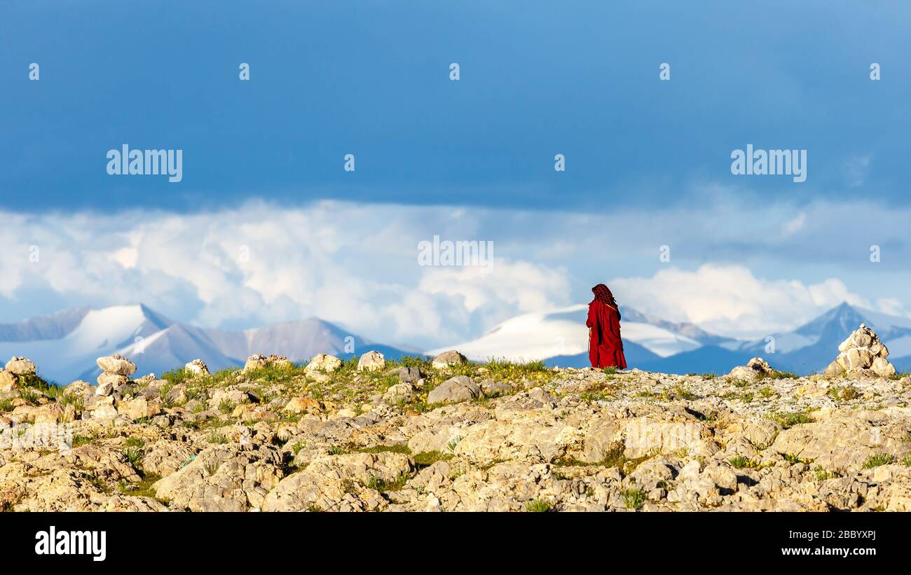 Donna, pellegrino buddista in veste rossa camminando nel deserto. Sullo sfondo montagne dell'altopiano tibetano. Vicino al lago Nam Co. Cielo drammatico Foto Stock
