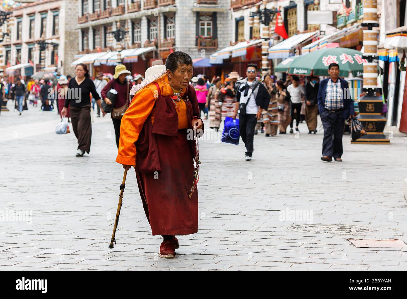 Anziani, pellegrino buddista in arancione e rosso con bastone da passeggio in via Barkhor. Sullo sfondo turisti e tipici edifici tibetani. Foto Stock