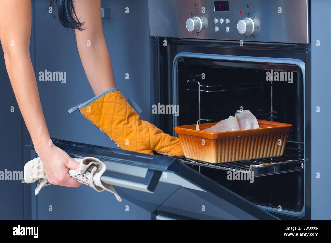 Tostatura in forno. Le mani femminili mettono un foglio di cottura all'interno del forno e lo chiudono. Sulla mano delle ragazze è un guanto della cucina. Foto Stock