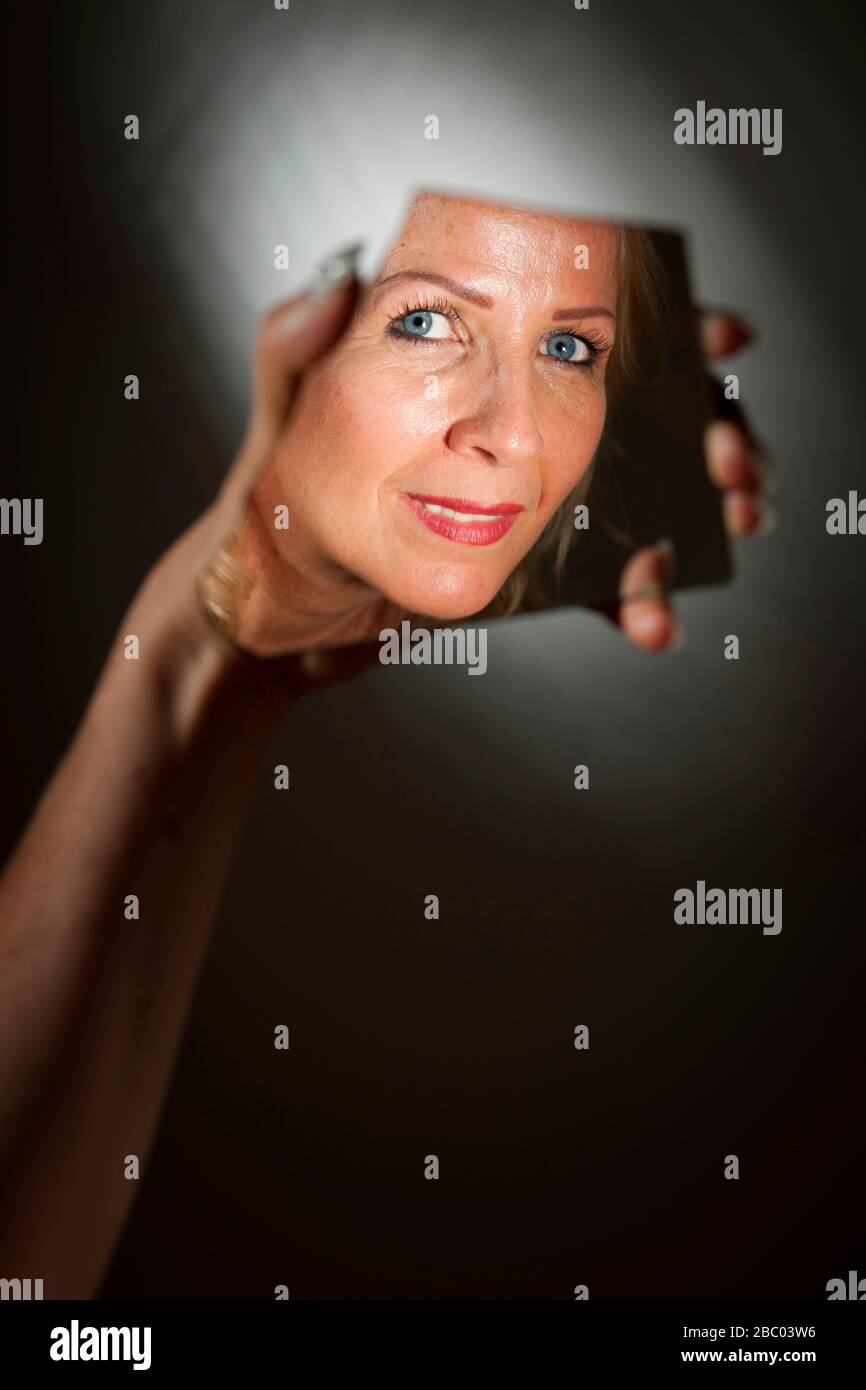 Immagine speculare del volto di una donna bionda di mezza età in uno specchio a mano brilla su uno sfondo nero con luce spot. Foto Stock