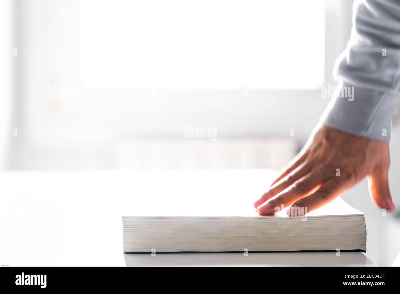 Chiusura di un libro con la mano su un tavolo bianco con la luce dalla finestra sullo sfondo Foto Stock