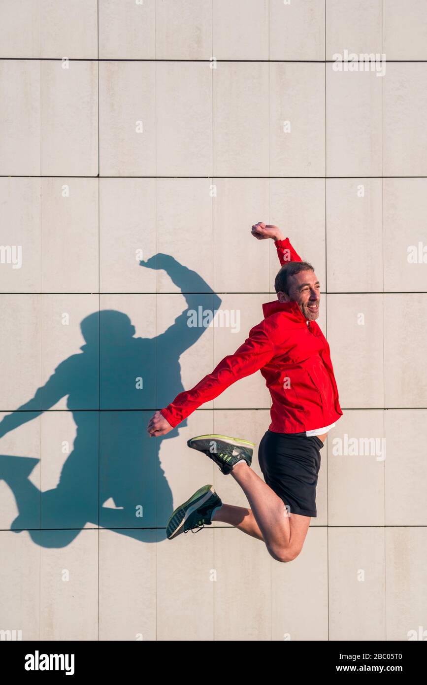 L'atleta caucasico indossa giacca rossa e pantaloni neri e salta con felicità in una giornata di sole con la sua ombra gettata su una parete bianca. Foto Stock