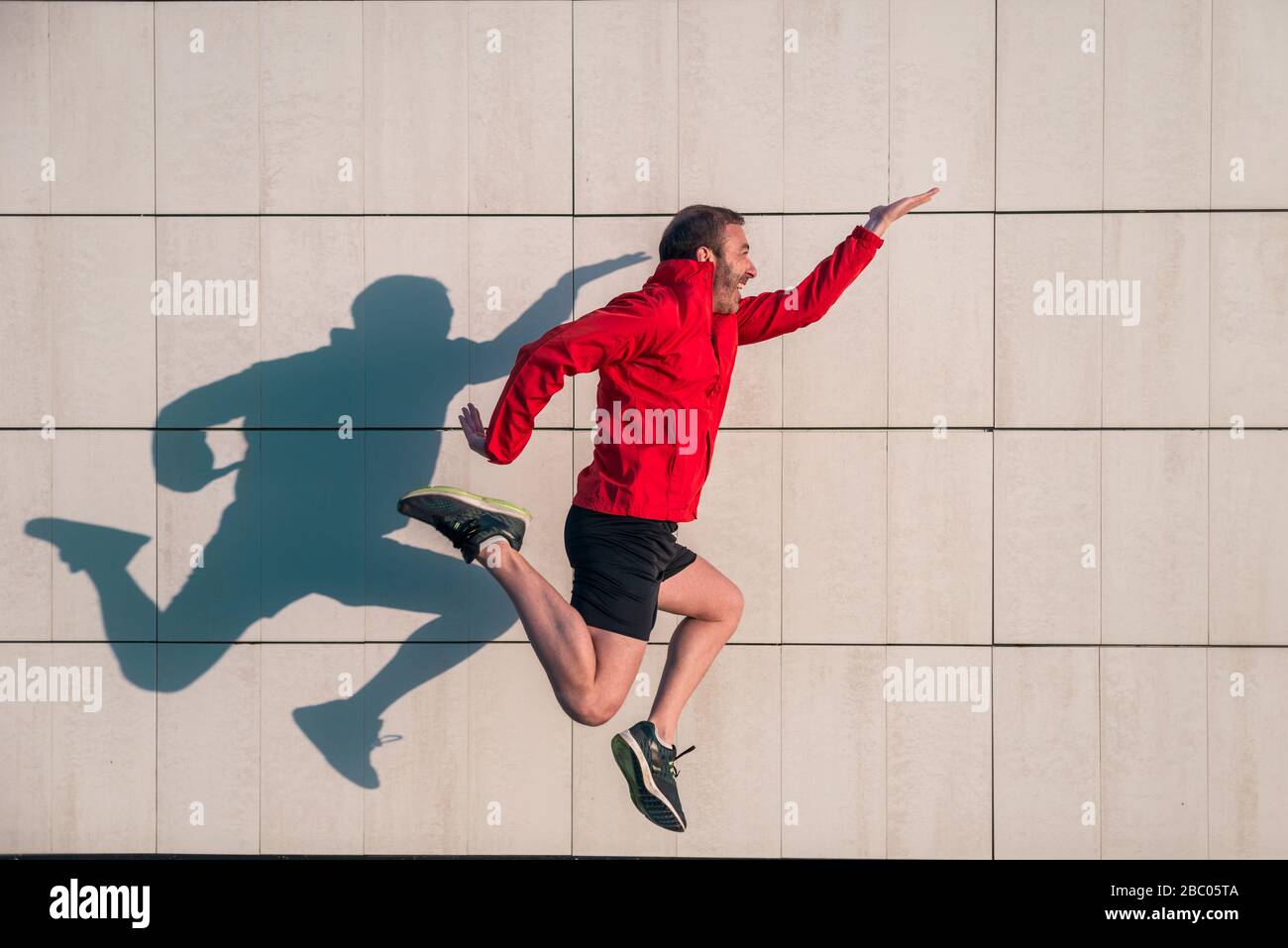 L'atleta caucasico indossa giacca rossa e pantaloni neri e salta con felicità in una giornata di sole con la sua ombra gettata su una parete bianca. Foto Stock