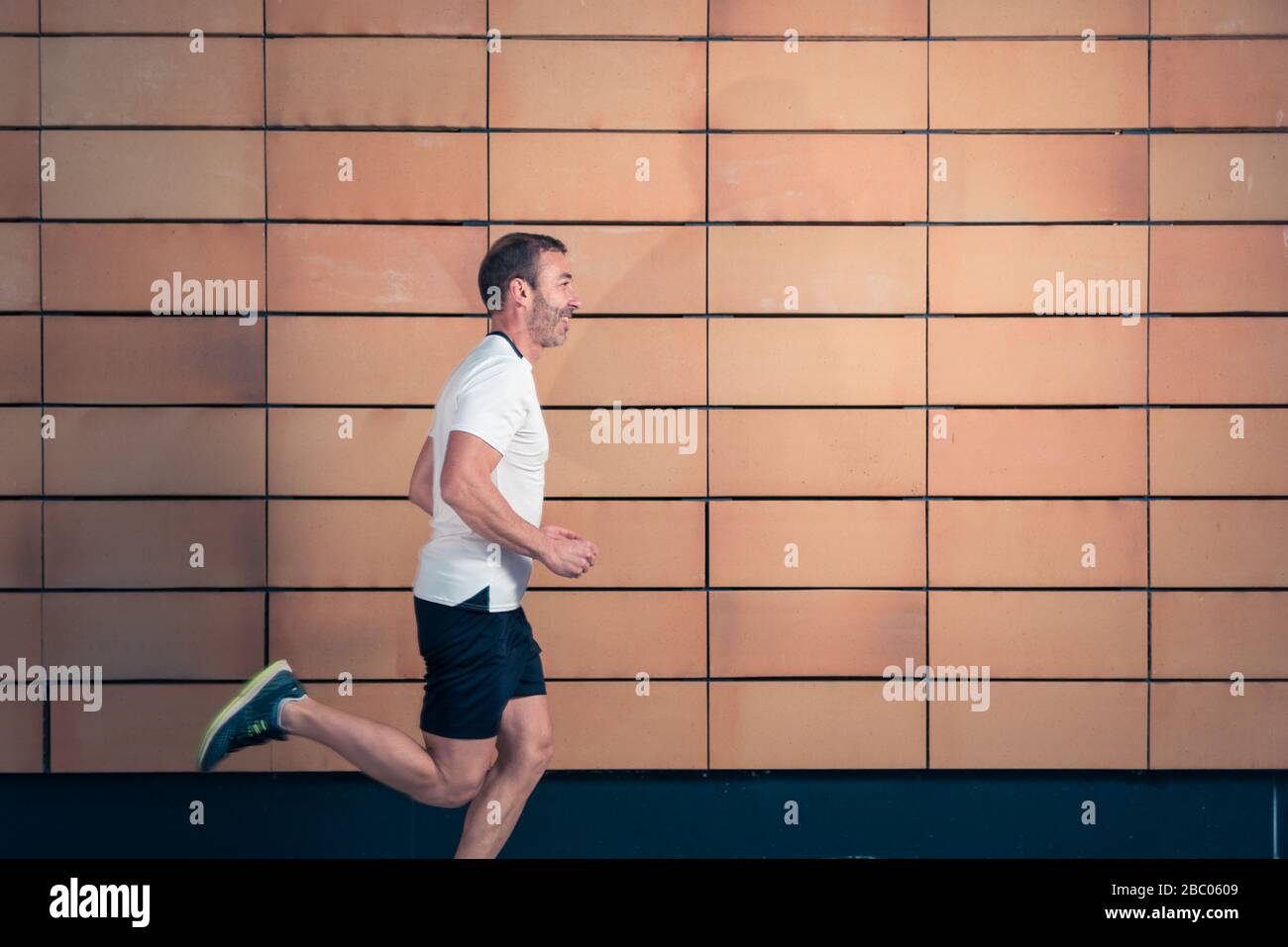 Atleta caucasico uomo in t-shirt bianca e pantaloncini neri, correndo in profilo accanto a un muro di mattoni arancioni. Foto Stock