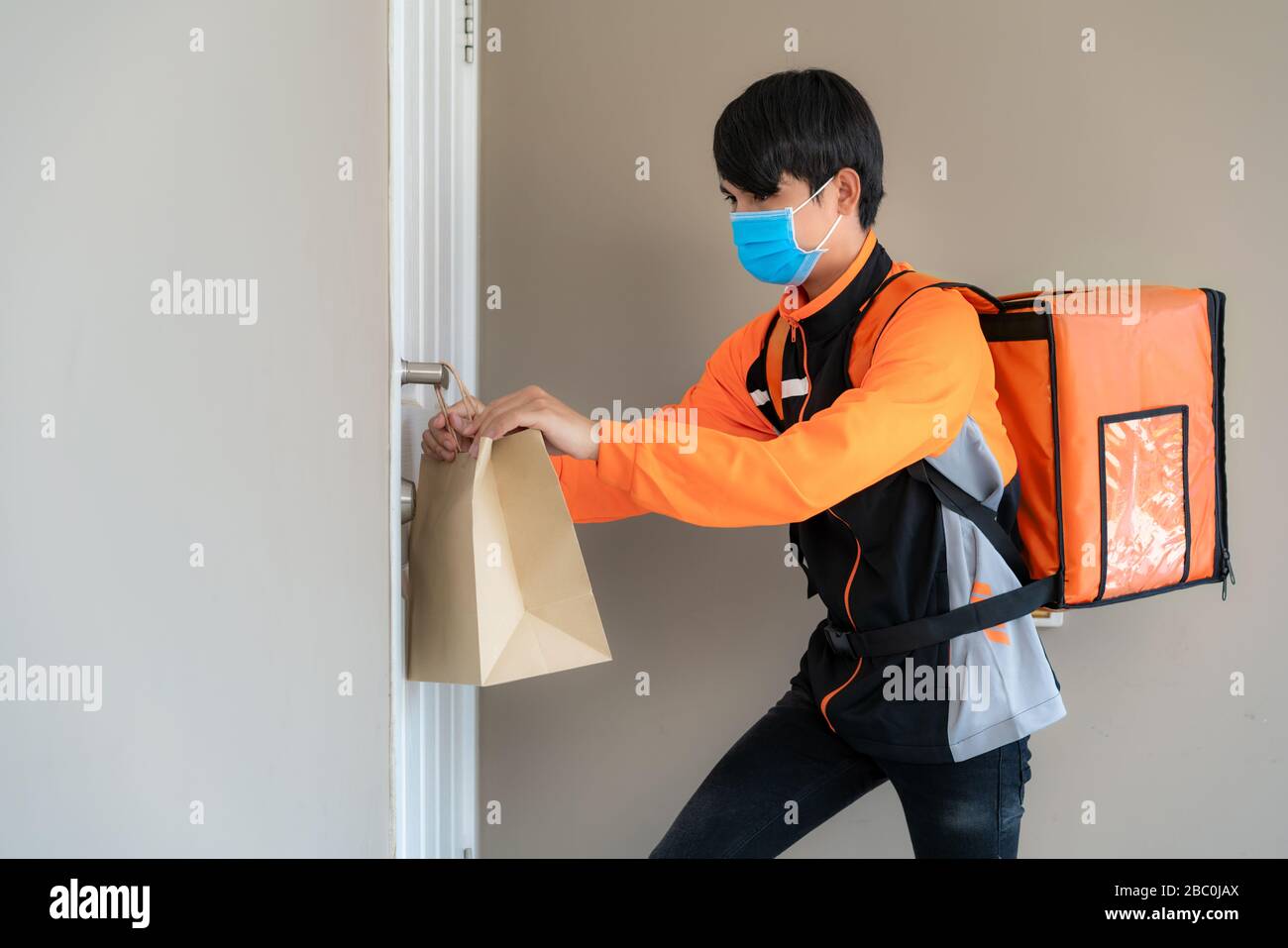 L'uomo di consegna asiatico invia il sacchetto del cibo alla manopola della porta per contactless o il contatto libero dal rider di consegna nella casa anteriore per distancing sociale per l'infezione r Foto Stock