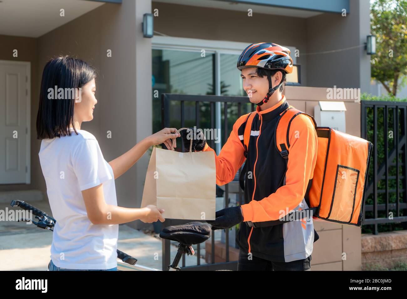 Asian uomo corriere sulla bicicletta che consegna il cibo nel sorriso uniforme arancione e che tiene la borsa del cibo nella casa di fronte e donna asiatica che accetta una consegna delle scatole Foto Stock