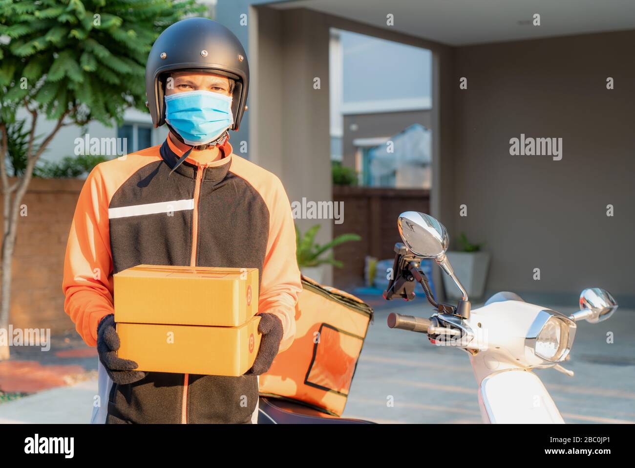 Asian consegna giovane uomo con maschera protettiva in uniforme arancione reggendo pile di scatole di cartone in casa villaggio di fronte con scooter. Pubblicità, Busi Foto Stock