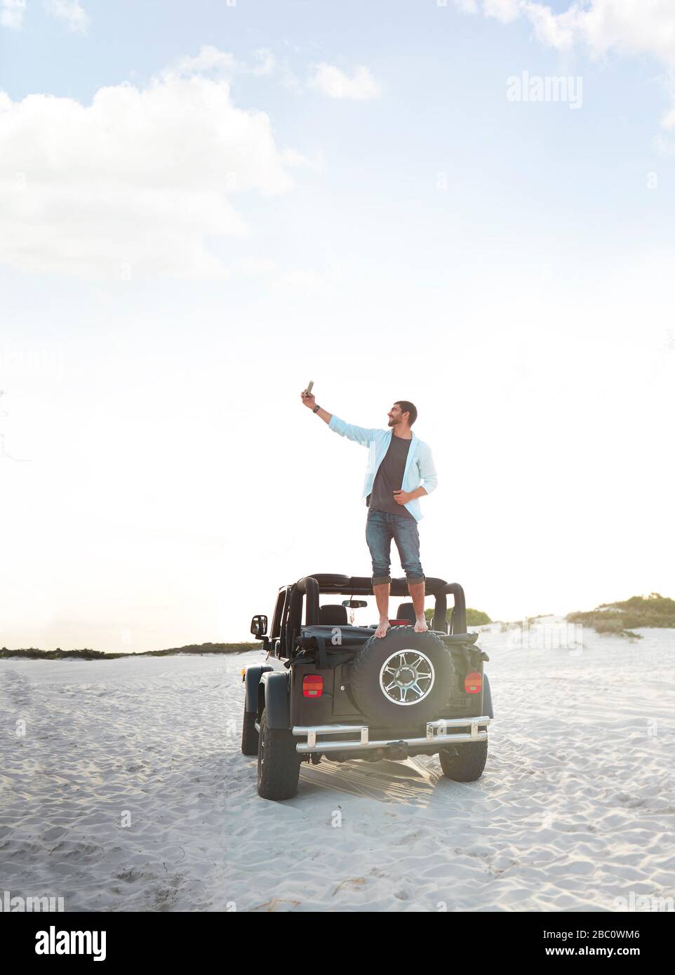 Giovane uomo con fotocamera telefono che prende selfie in cima a jeep pneumatico sulla spiaggia Foto Stock