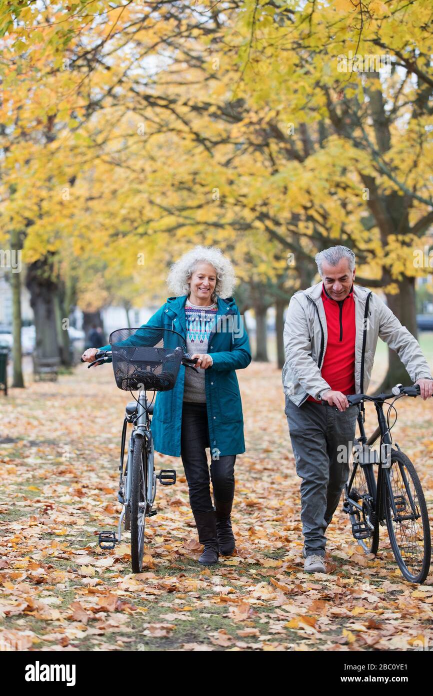 Coppia senior biciclette a piedi tra alberi e foglie nel parco autunnale Foto Stock