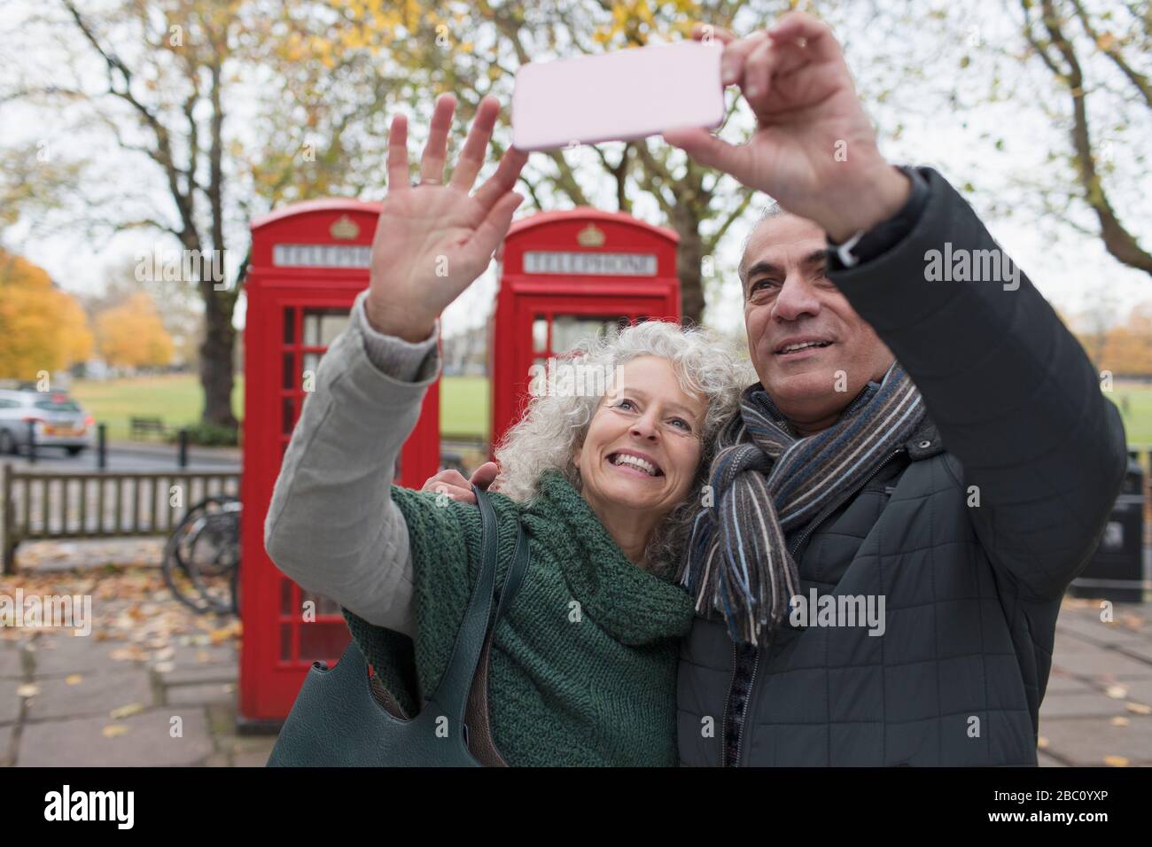 Sorridente coppia senior con selfie nel parco autunnale di fronte alle cabine telefoniche rosse Foto Stock