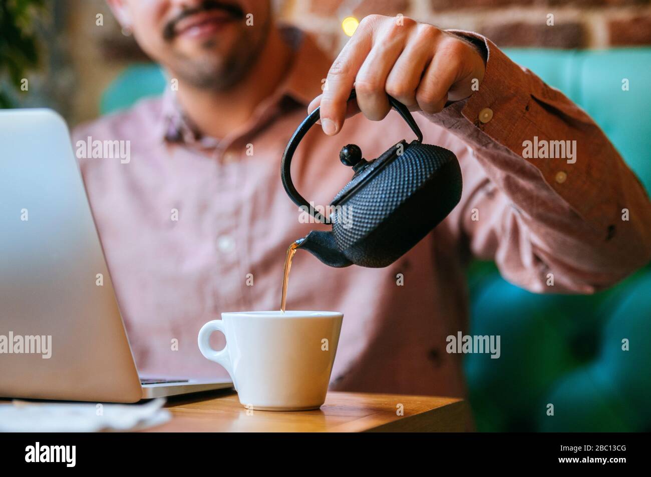 Uomo con computer portatile in un caffè che versa il tè in una tazza Foto Stock
