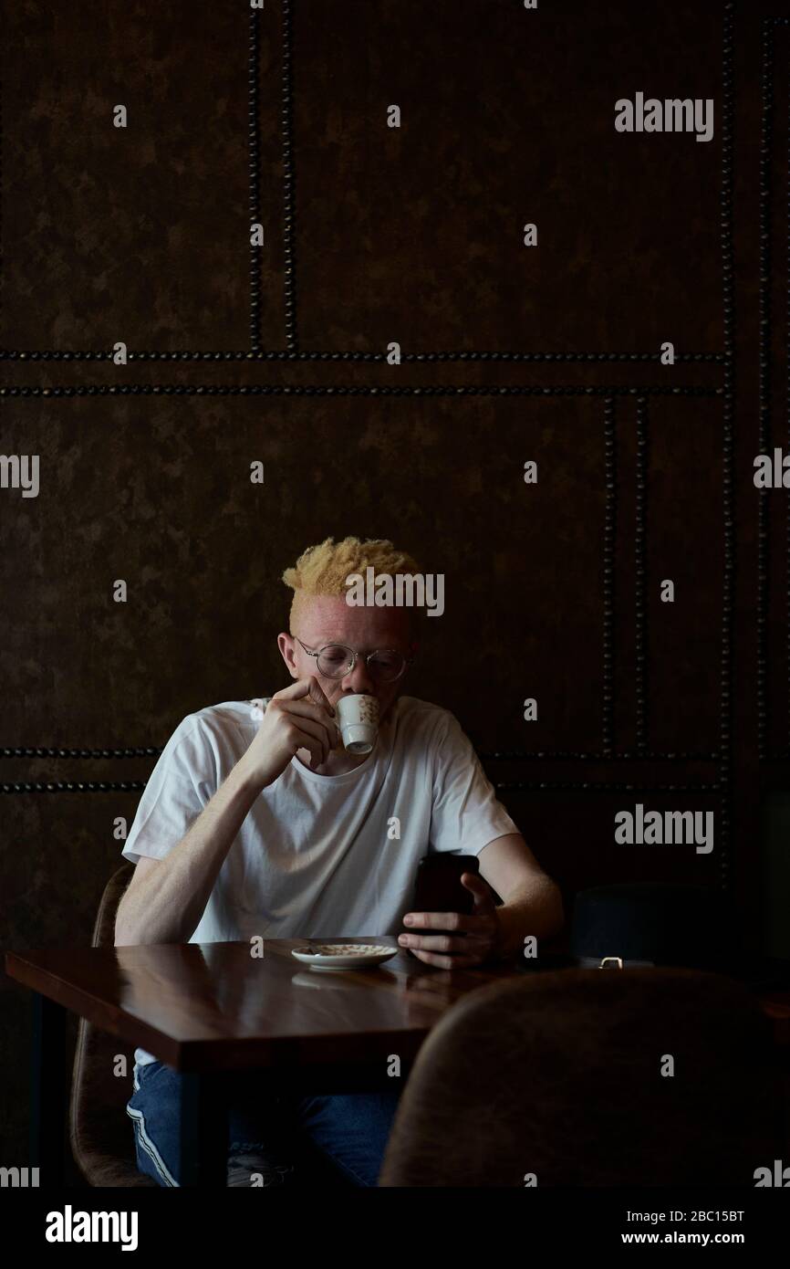 Albino uomo con bicchieri rotondi che ha un caffè e che controlla il suo telefono in una caffetteria Foto Stock