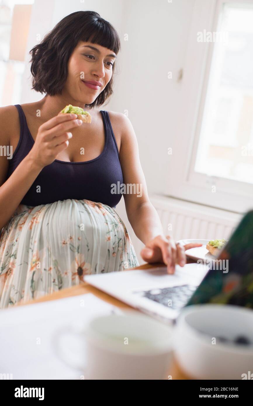 Donna incinta che mangia al computer portatile Foto Stock
