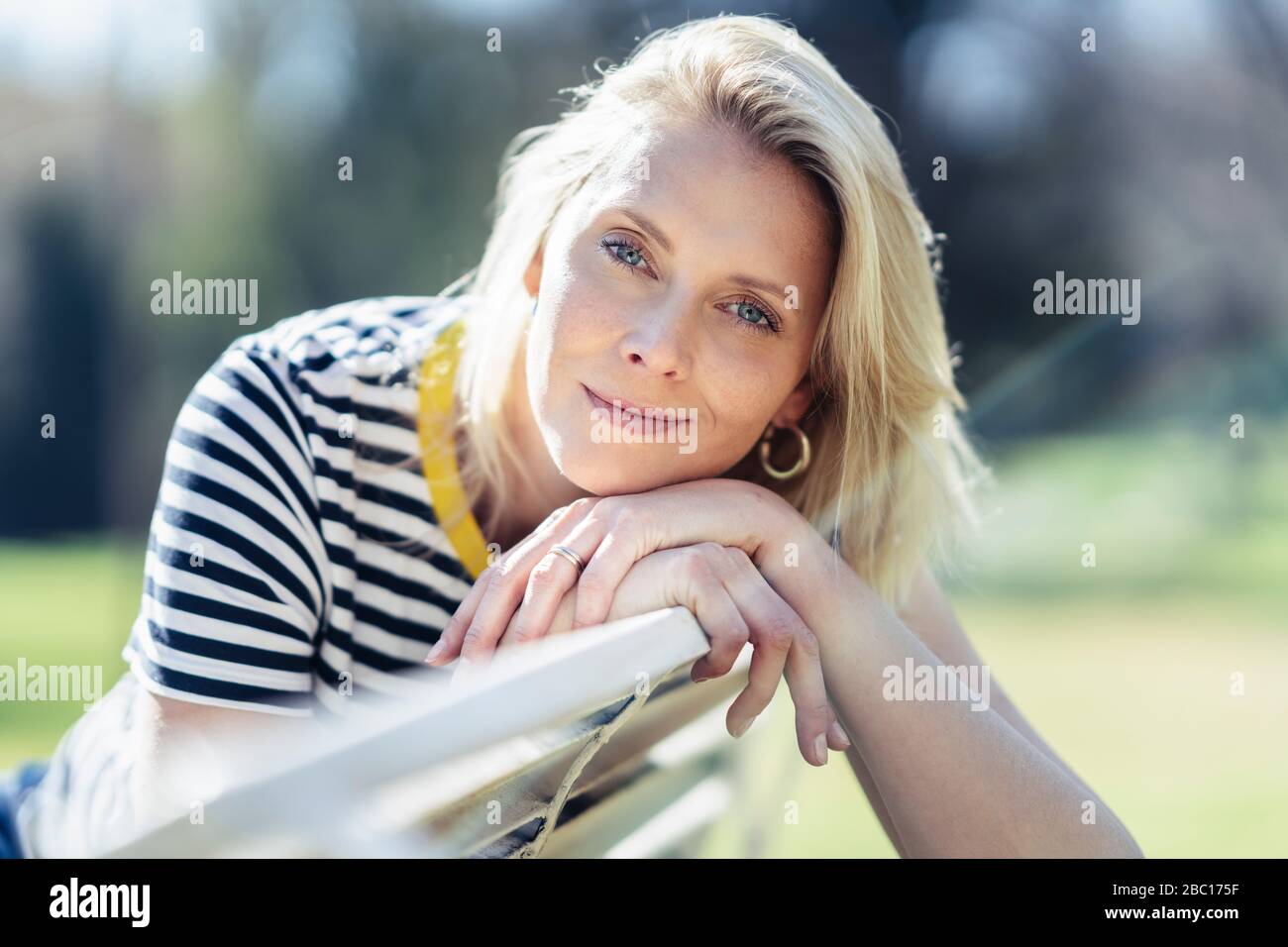 Donna bionda sorridente seduta su una panchina e guardando la macchina fotografica Foto Stock