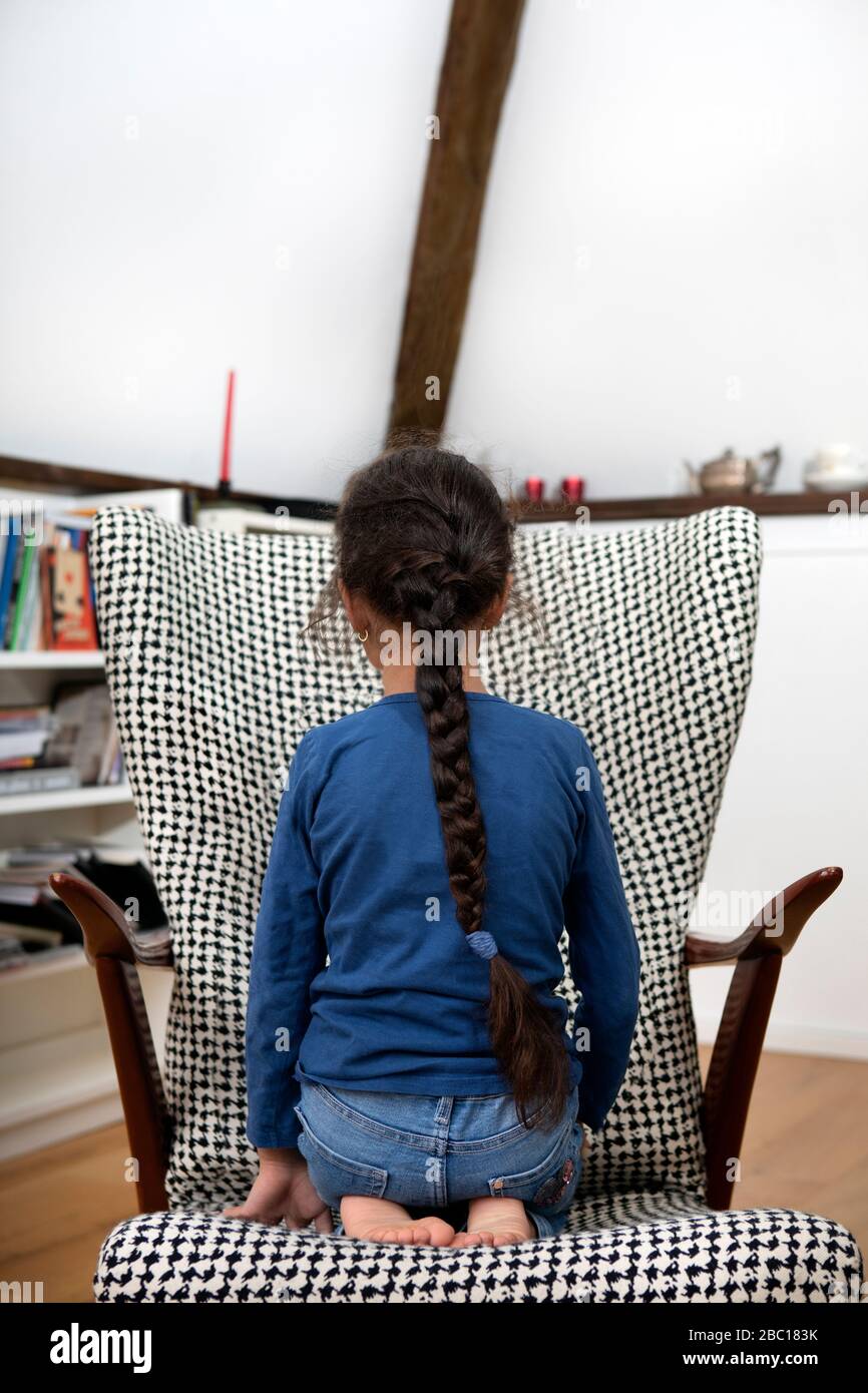 Vista posteriore della bambina con treccia lunga accovacciata su una poltrona Foto Stock