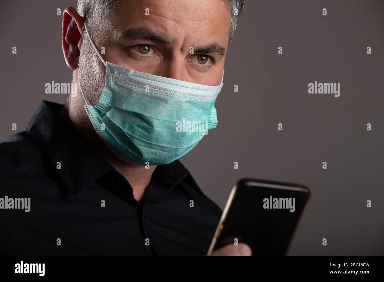 Uomo con maschera di protezione chirurgica con volto preoccupato dopo aver guardato le notizie sul cellulare su sfondo grigio. Concetto di coronavirus. Proteggere la salute. Foto Stock
