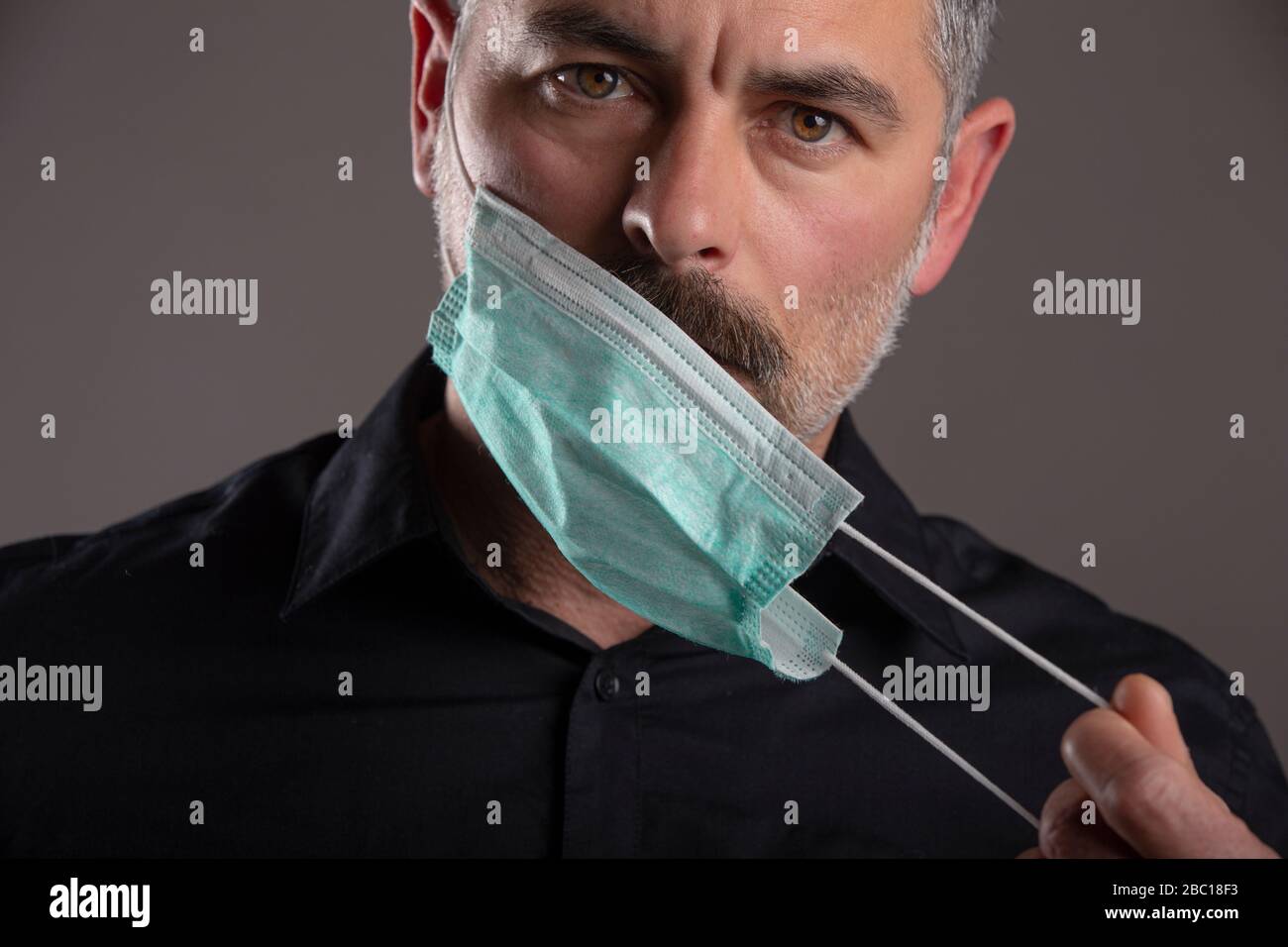 Ritratto di atteggiamento serio uomo togliere maschera di protezione medica su sfondo grigio studio. Concetto di responsabilità personale durante la quarantena. Foto Stock