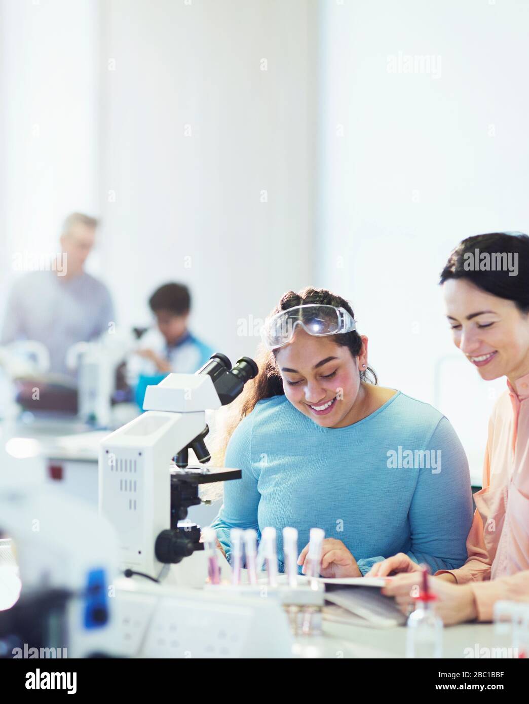 Sorridente insegnante di scienza femminile e studentessa studentessa che legge il libro di testo al microscopio in classe di laboratorio Foto Stock