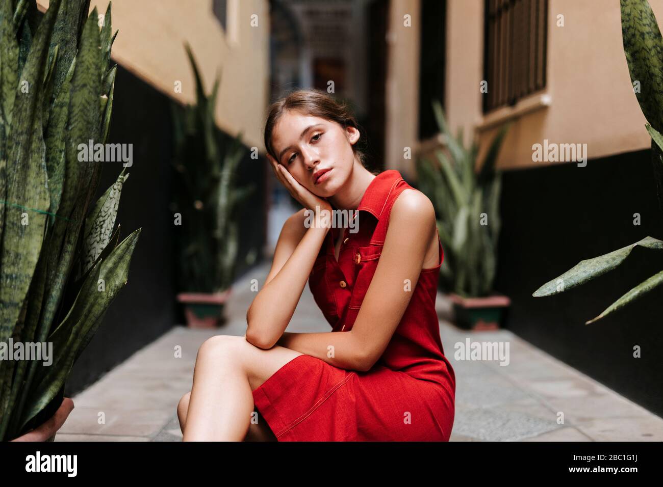 Ritratto di giovane donna malinconica con abito rosso seduto sul pavimento urbano, a piedi nudi e circondato da vasi verdi a Siviglia, Spagna Foto Stock