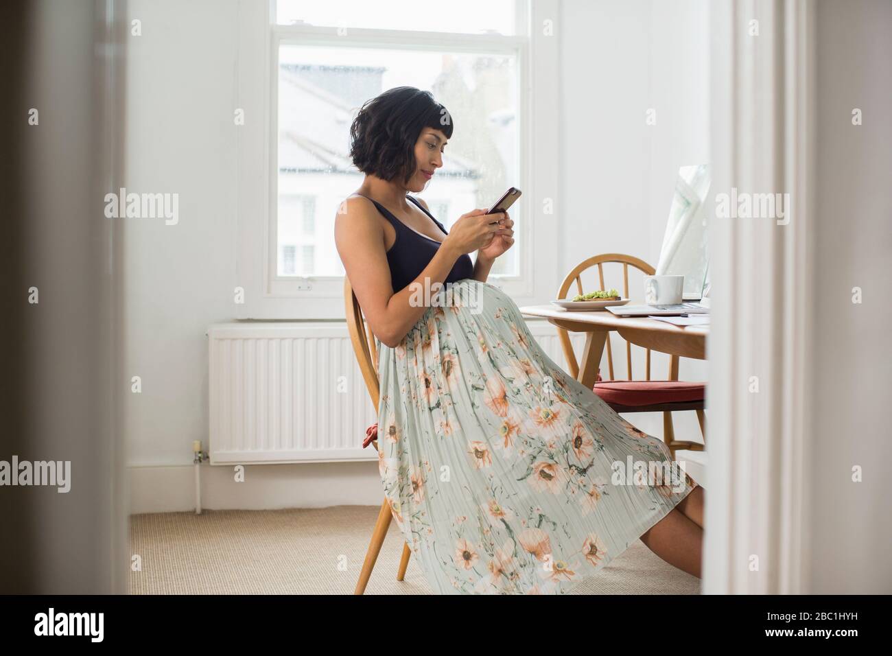 Donna incinta in abito floreale con smartphone al tavolo Foto Stock