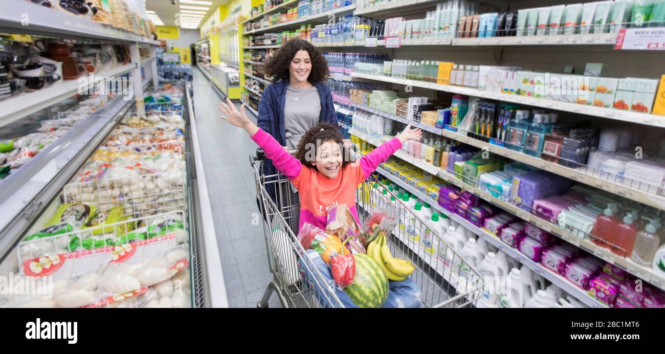 Madre che spinge la figlia giocosa nel carrello della spesa in corridoio del supermercato Foto Stock
