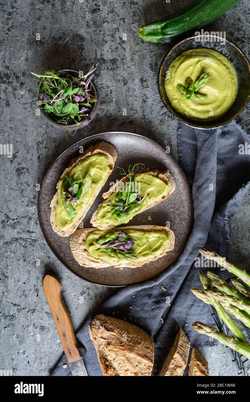 Fette di pane con avocado vegetariano, zucchine e asparagi si spargero, sormontate da piselli e germogli di ravanello su un piatto di ceramica Foto Stock