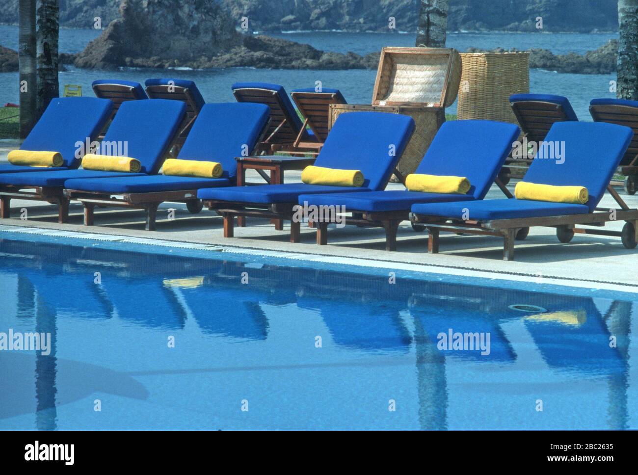 Fila di sedie a sdraio blu con asciugamani gialli allineati lungo il lato di una piscina in un hotel a Careyes, Jalisco, Messico. Essi sono riflessi nell'acqua. Foto Stock