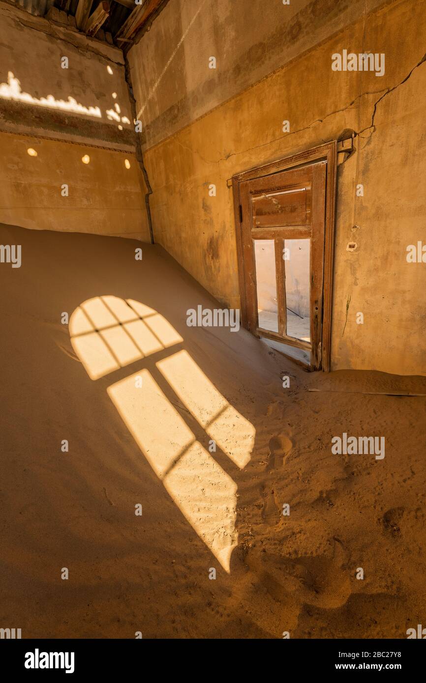 Una fotografia verticale all'interno di una casa abbandonata con sabbia desertica accatastata nell'angolo e luce solare dorata che scorre attraverso una porta rotta, presa nel Foto Stock