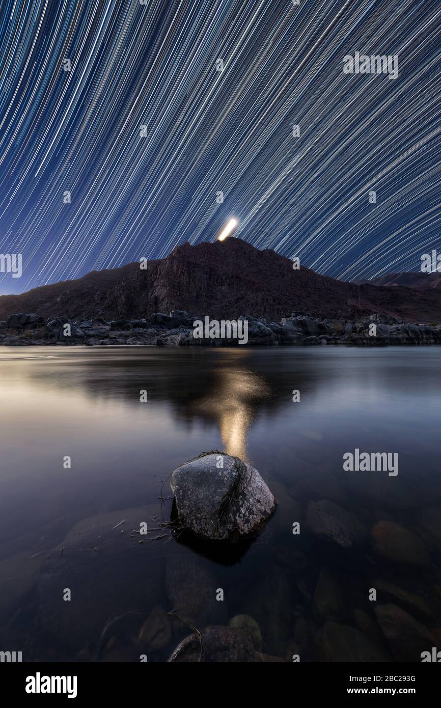 Un bellissimo paesaggio verticale notturno con sentieri stellari contro un cielo blu profondo e la luna dietro le montagne, che si riflette nella Rive arancione Foto Stock