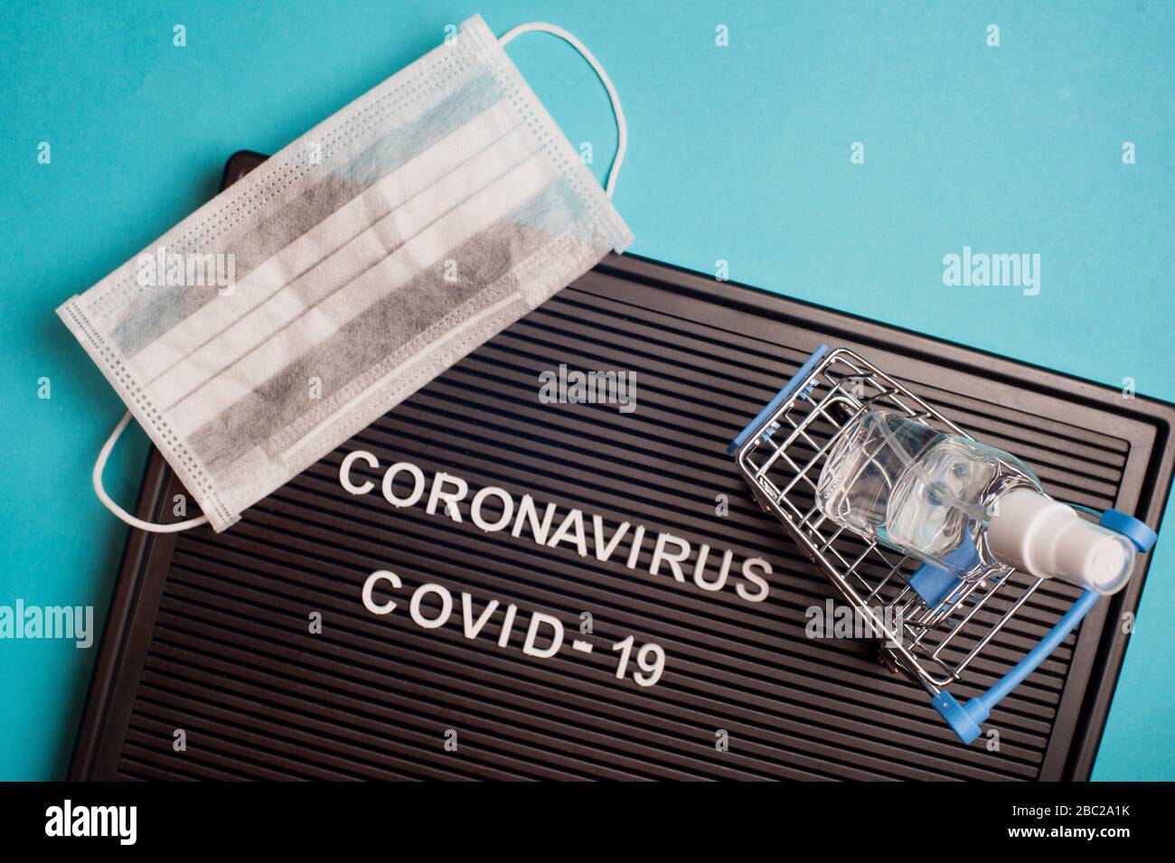 Coronavirus - COVID -19 - testo su bordo nero della lettera, maschera chirurgica e gel igienizzante in piccolo carrello su sfondo blu. Foto Stock