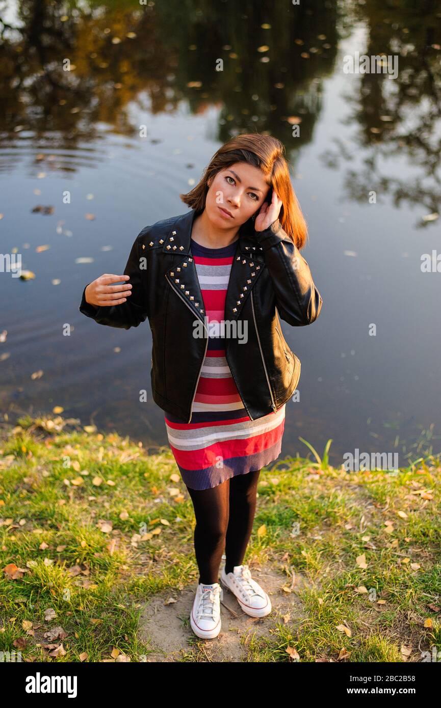 bella ragazza adolescente in sneakers bianche, un abito a maglia a righe  rosse e una giacca nera, raddrizza i capelli scuri, in piedi accanto al  fiume Foto stock - Alamy