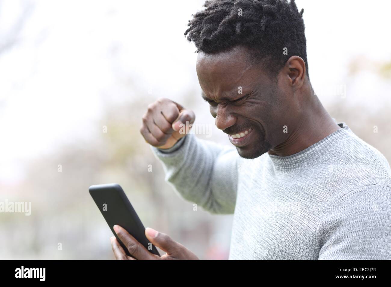 Uomo nero arrabbiato che controlla i contenuti su smartphone con pugno chiuso in strada Foto Stock