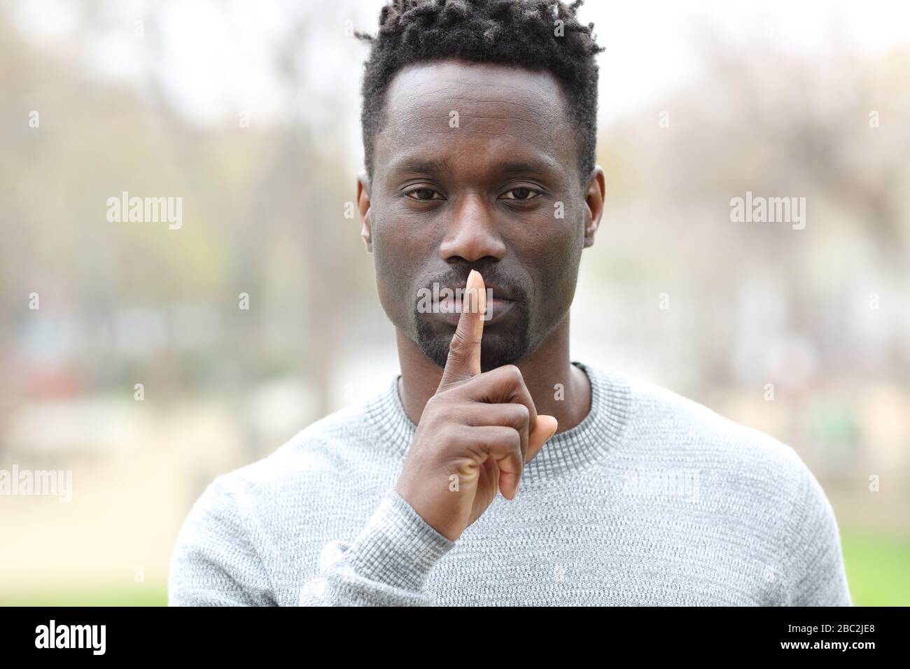 Vista frontale ritratto di un uomo nero che chiede silenzio con un dito sulle labbra in piedi in un parco Foto Stock
