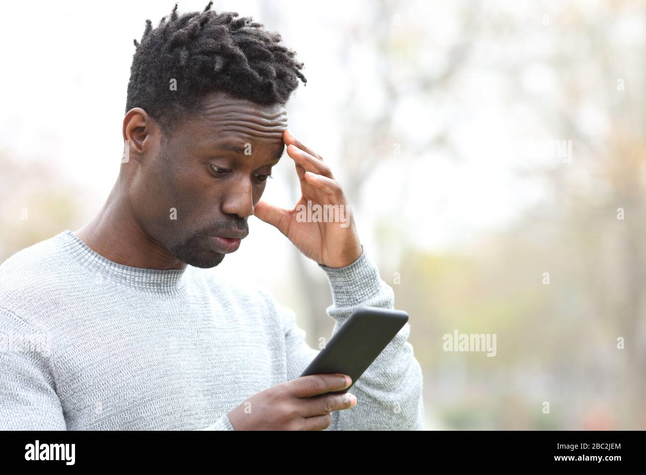 L'uomo nero si lamenta dopo aver commesso un errore su uno smartphone in piedi in un parco Foto Stock