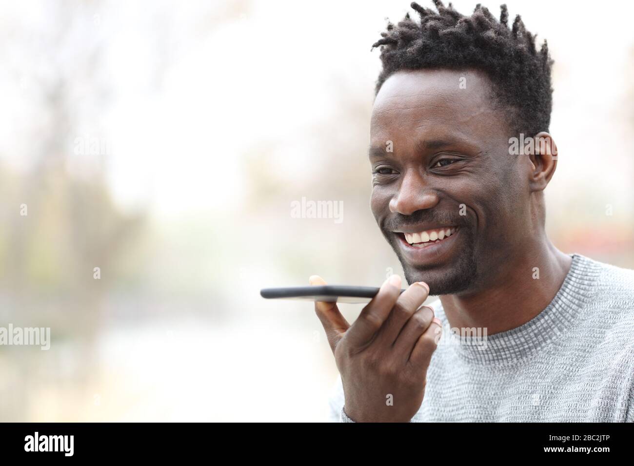 Uomo nero felice che utilizza il riconoscimento vocale su smartphone all'aperto in un parco Foto Stock
