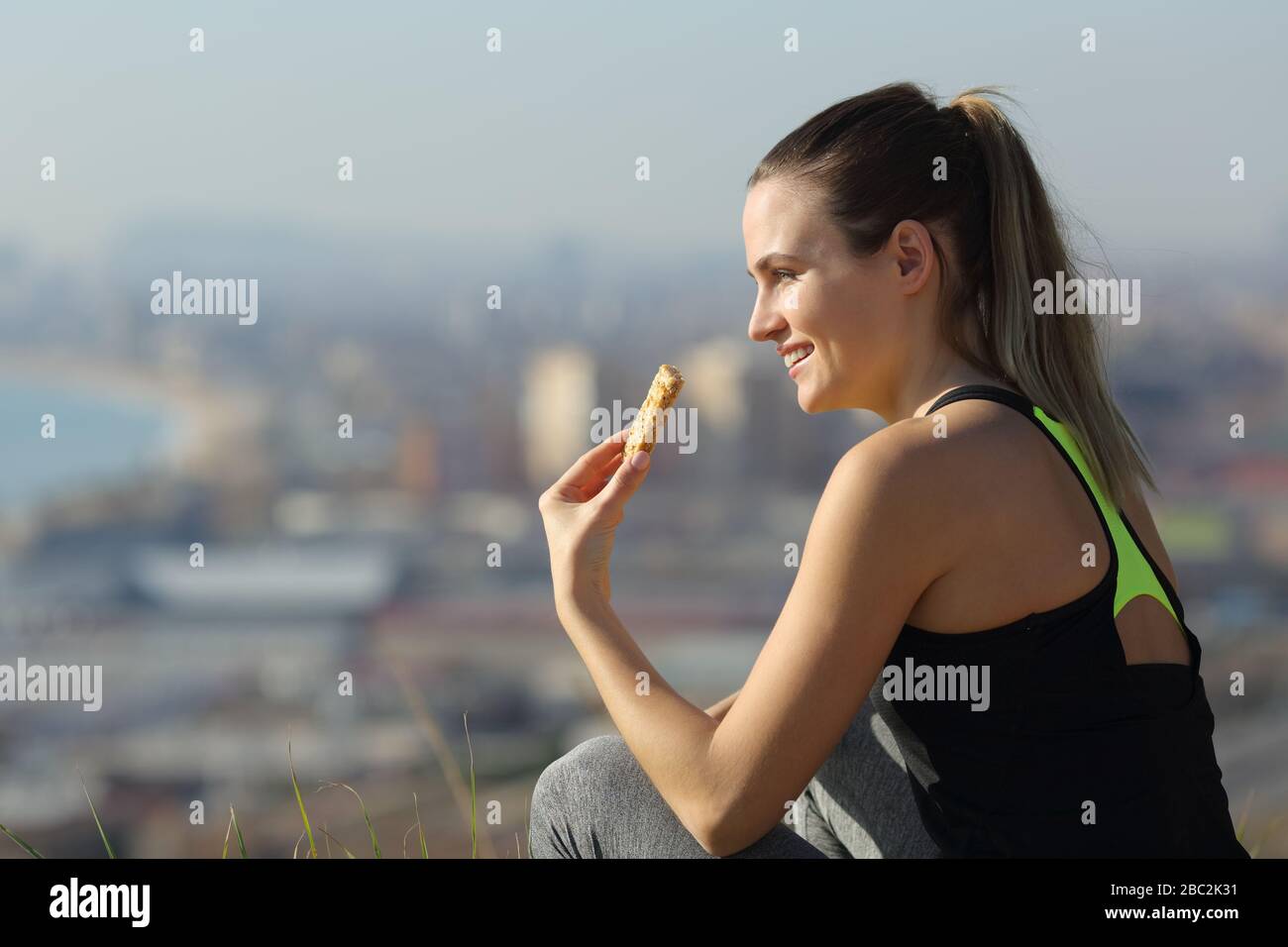 Donna runner felice mangiare bar energia seduti all'aperto dopo l'esercizio nella periferia della città Foto Stock