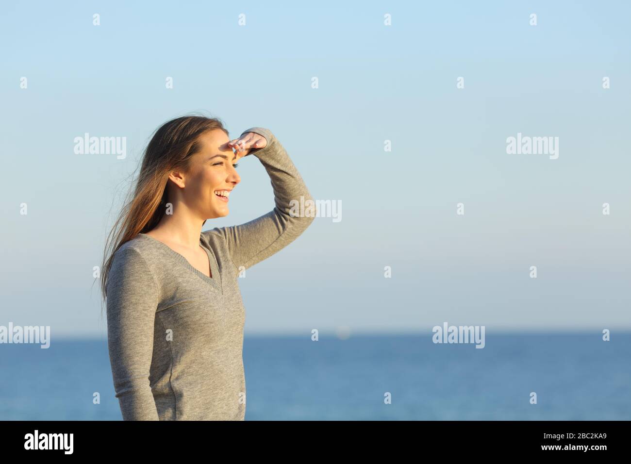 Donna felice che cerca guardando l'orizzonte sulla spiaggia con mano sulla fronte Foto Stock