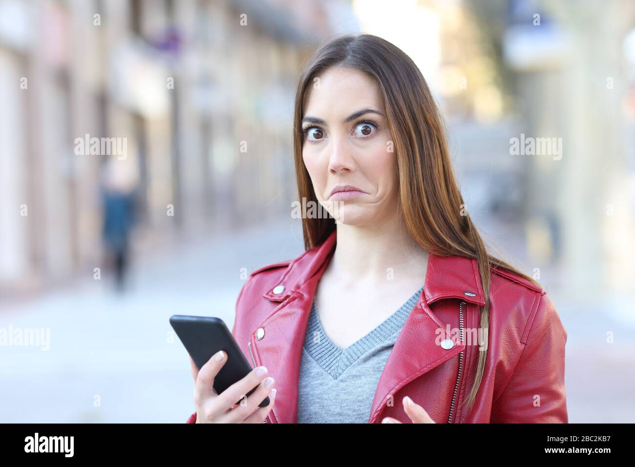 La donna perplessa che usa il telefono cellulare guarda la fotocamera in piedi in strada Foto Stock