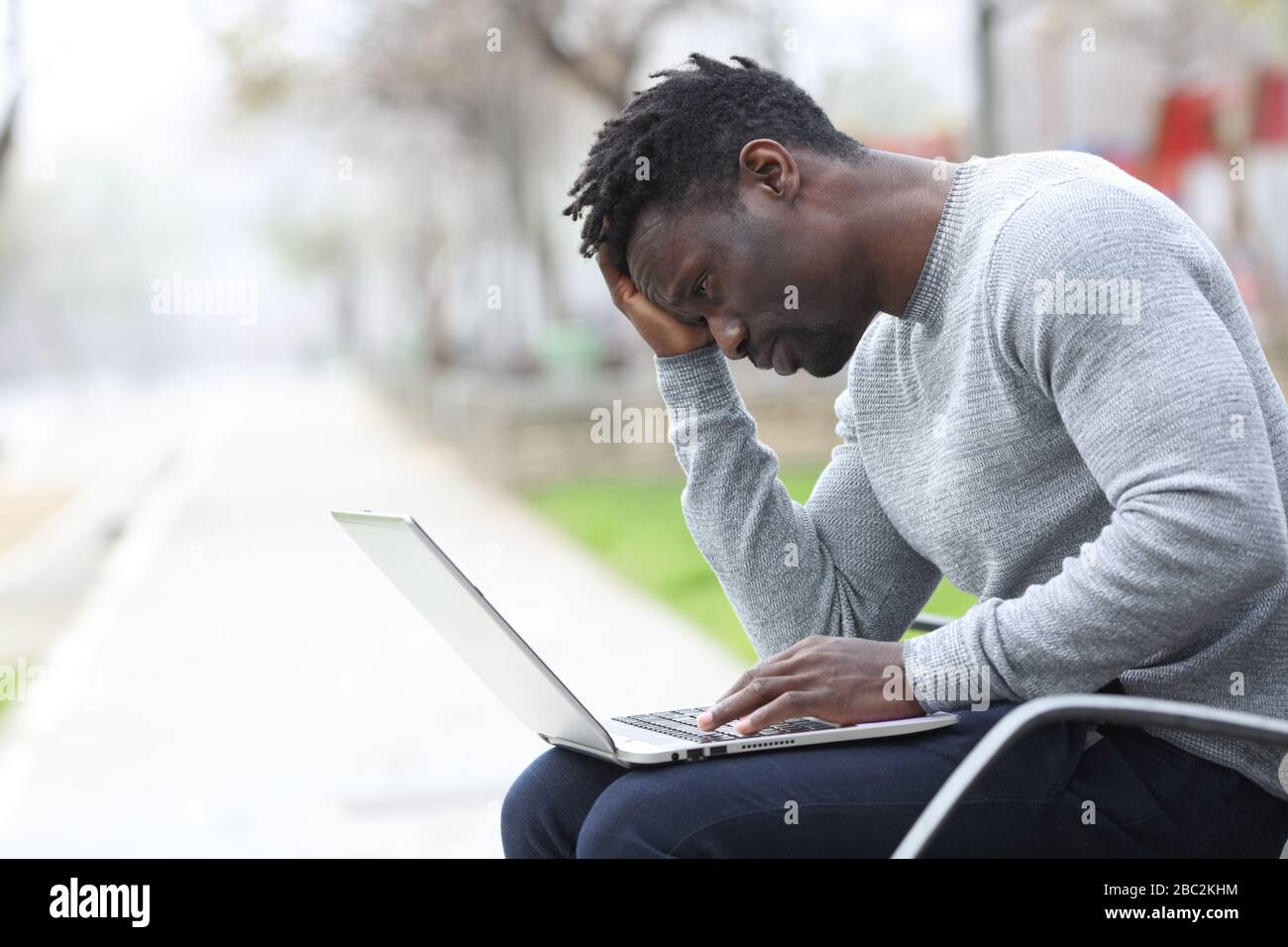 Triste uomo nero lamentarsi controllare il contenuto del laptop seduto su una panchina in un parco Foto Stock